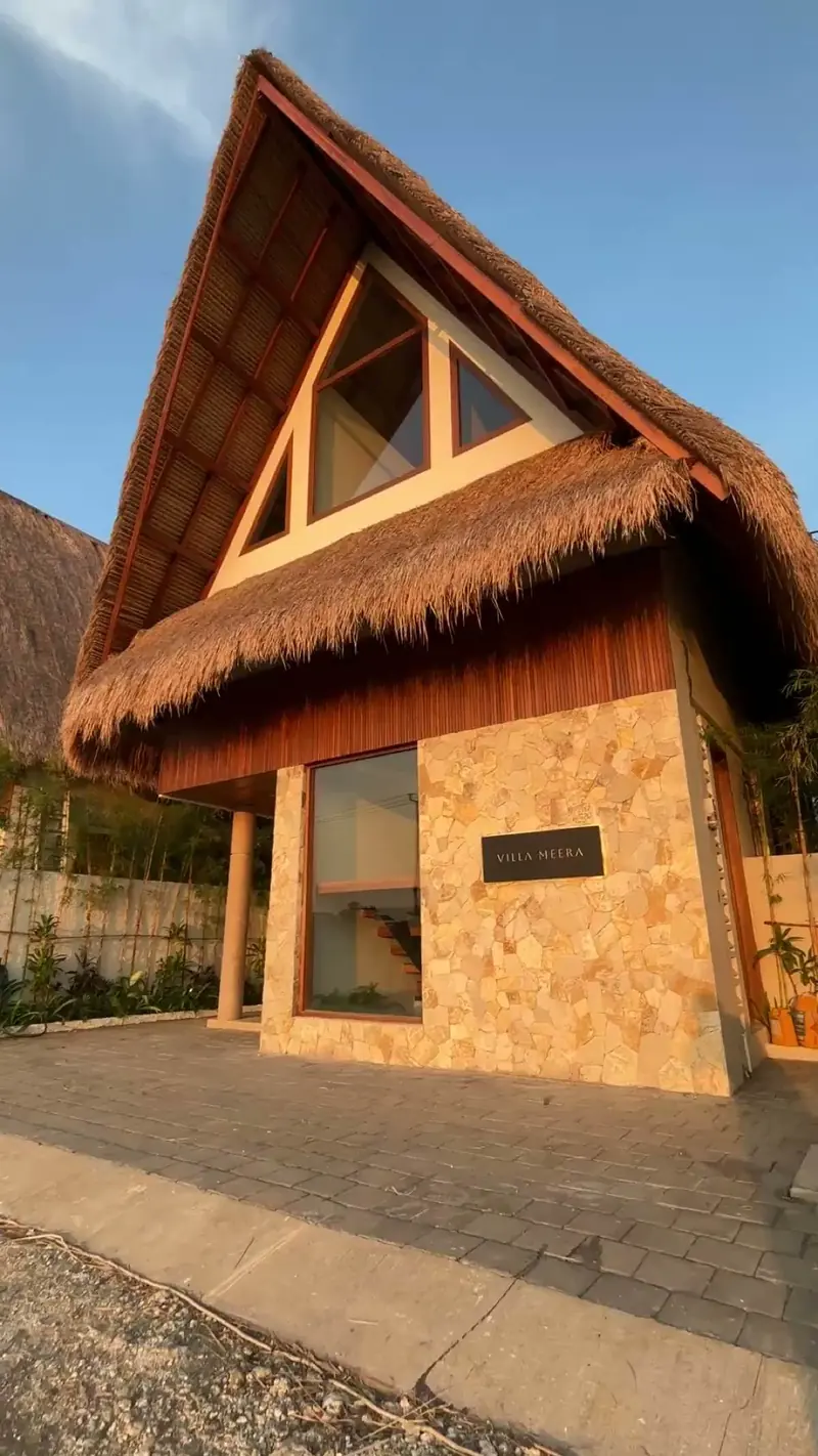 Photograph: the stone-clad entrance of a finished hut under the thatch, with a dark name plaque beside the door
