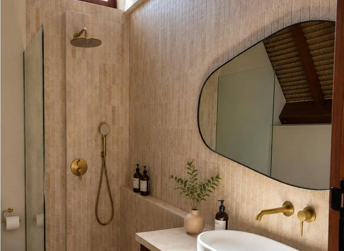 A bathroom with a white vessel basin on a stone counter, brass fittings and an irregular mirror against a textured wall