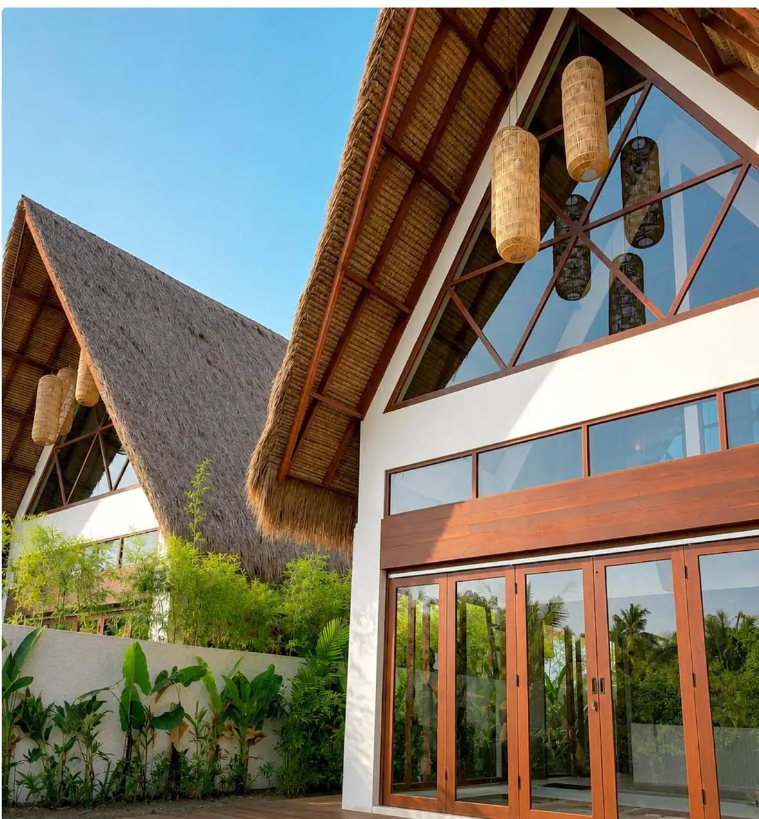 The gable end of an A-frame villa: hand-laid thatch sweeping to the ground, a triangular timber-framed window above and folding glass doors below